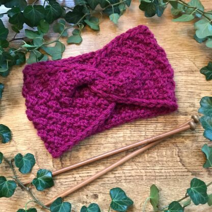 A deep pink knitted Aspen headband surrounded by ivy lies next to a pair of wooden knitting needles