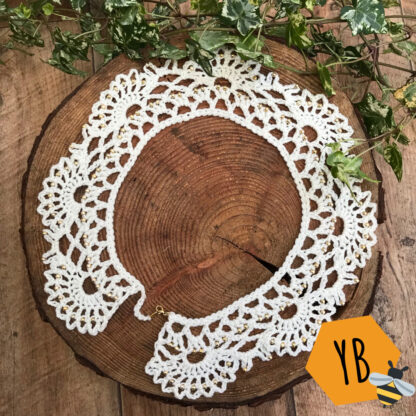 A cream lacy vintage style collar, with tiny decorative gold beads lies on a wood slice next to some ivy. The collar has a gold lobster clasp to fasten.